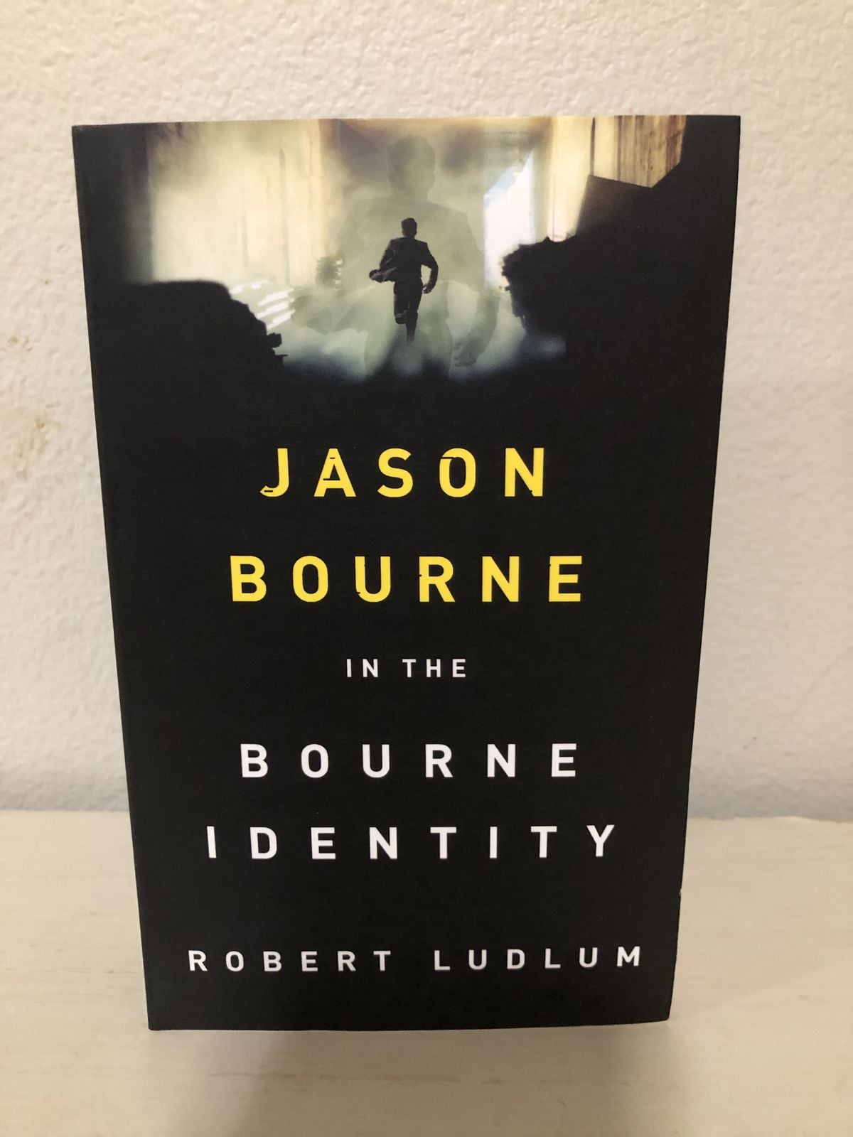 The cover of "The Bourne Identity" by Robert Ludlum features a silhouette of a running man in a foggy, dimly lit setting, with the title and author’s name in bold yellow and white text.