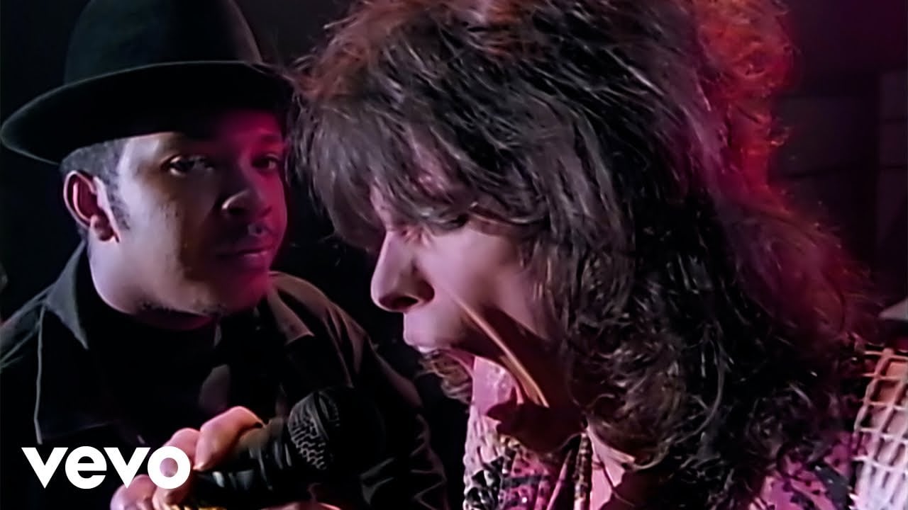 Two men performing on stage; one holds a microphone while the other energetically sings into it. The background is dark with hints of colorful lighting. The Vevo logo appears in the lower left corner.