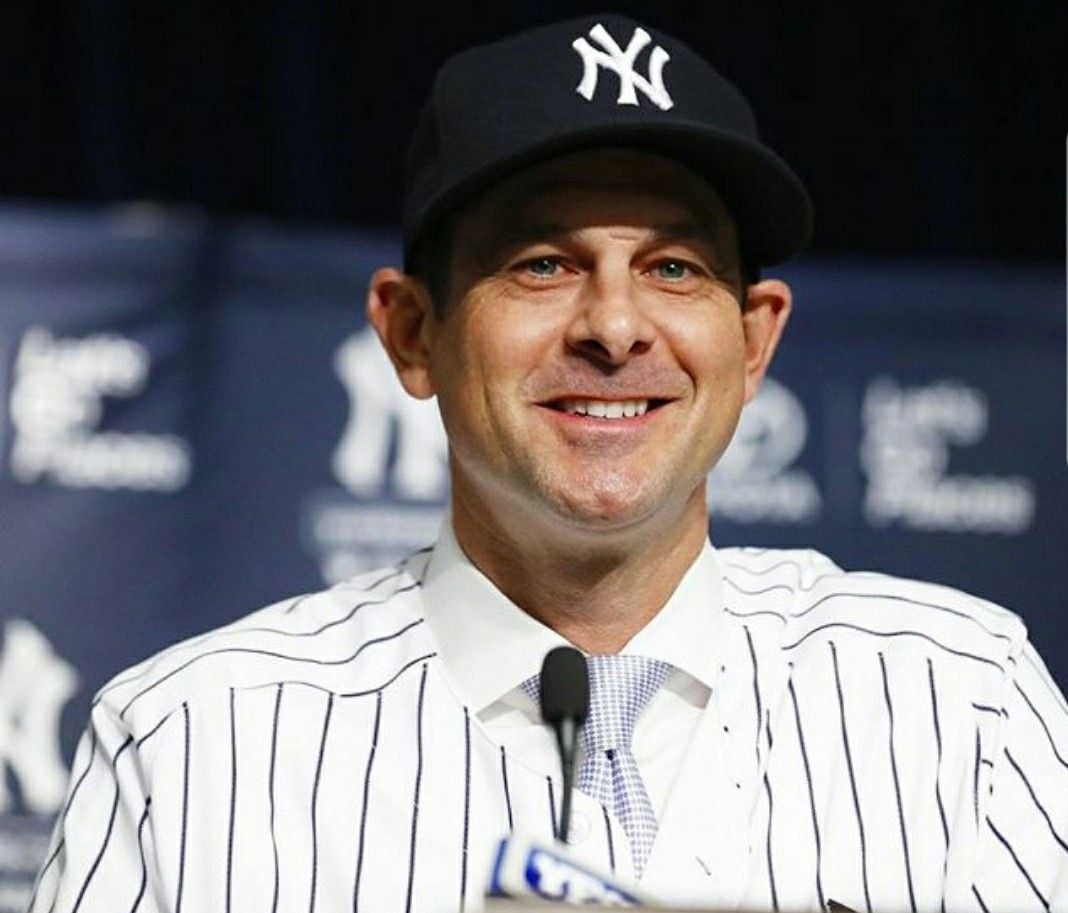 A man wearing a New York Yankees cap and pinstripe jersey smiles while sitting at a press conference table with a Yankees logo backdrop behind him.