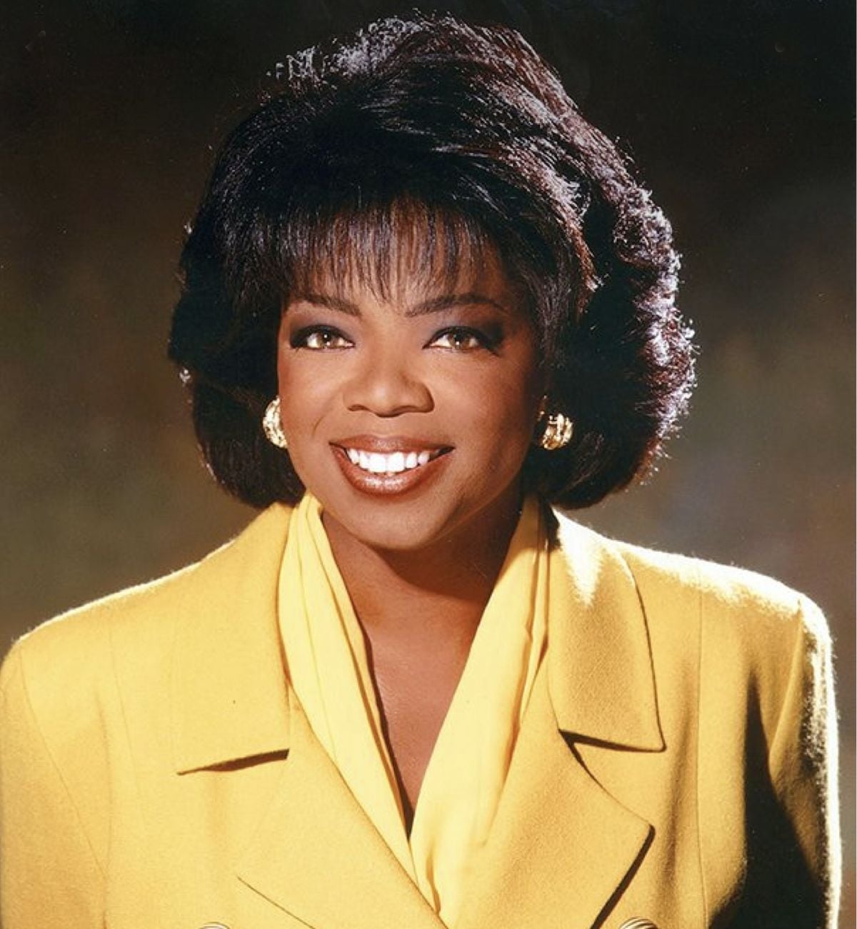 A woman with short, dark hair smiles warmly. She is wearing gold hoop earrings and a bright yellow blazer over a matching top. The background is softly lit in warm brown tones.
