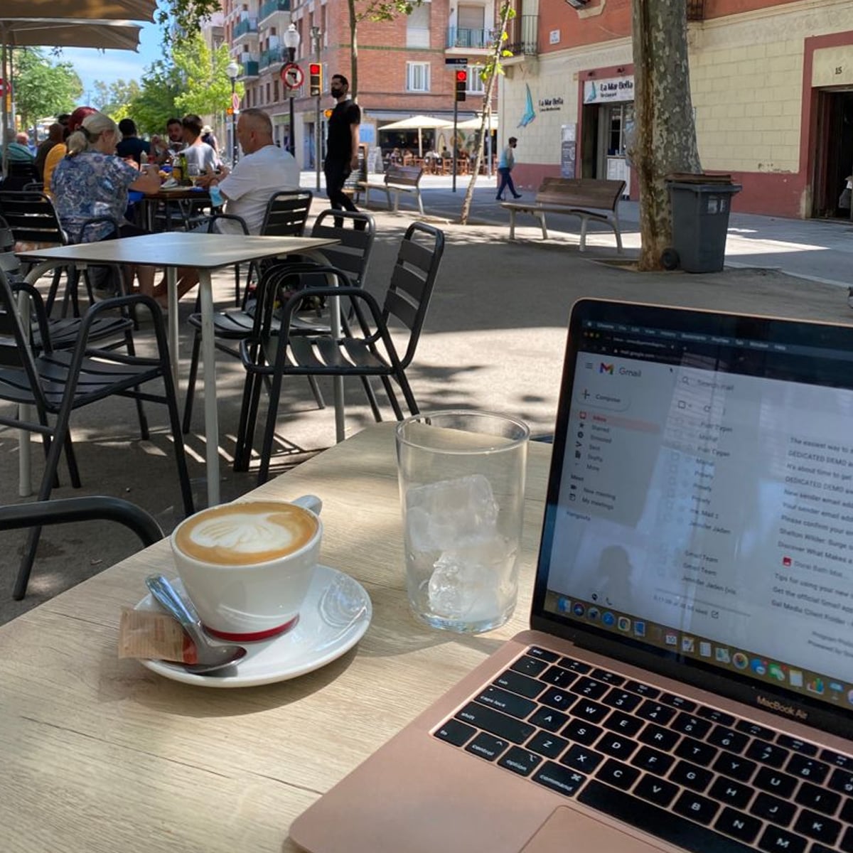 A laptop, a coffee cup with saucer, and a glass with ice sit on an outdoor cafe table; people are seated at other tables, and a sunny street with shops and trees is in the background.