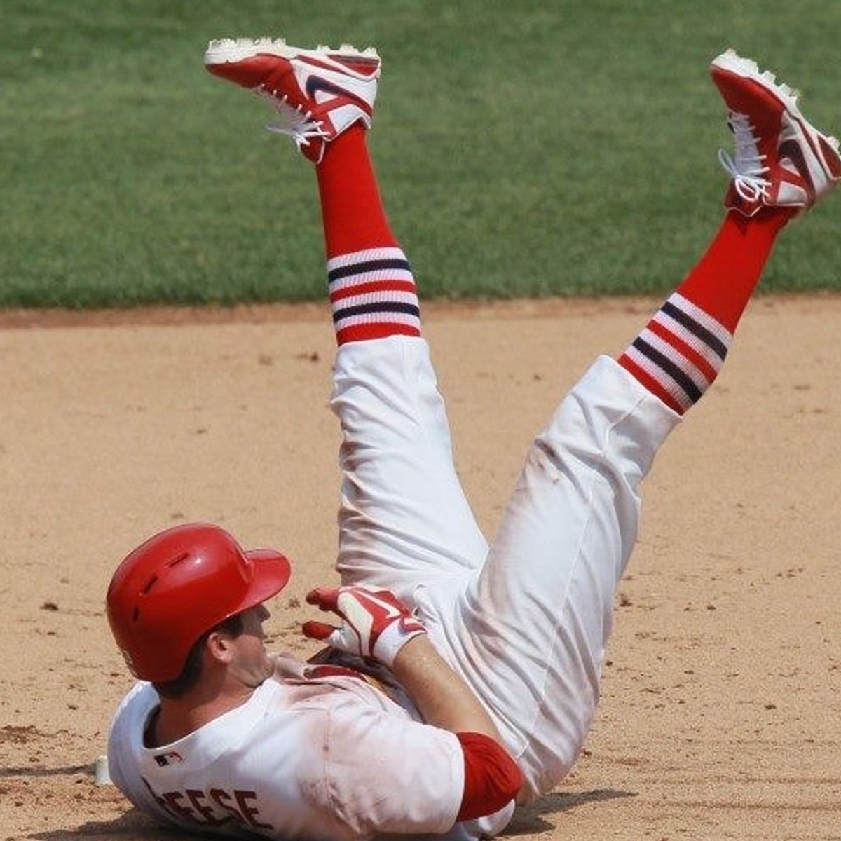 A baseball player in a red helmet and red-striped socks lies on his back on the field with one leg raised in the air, appearing to have just slid or fallen during a game.