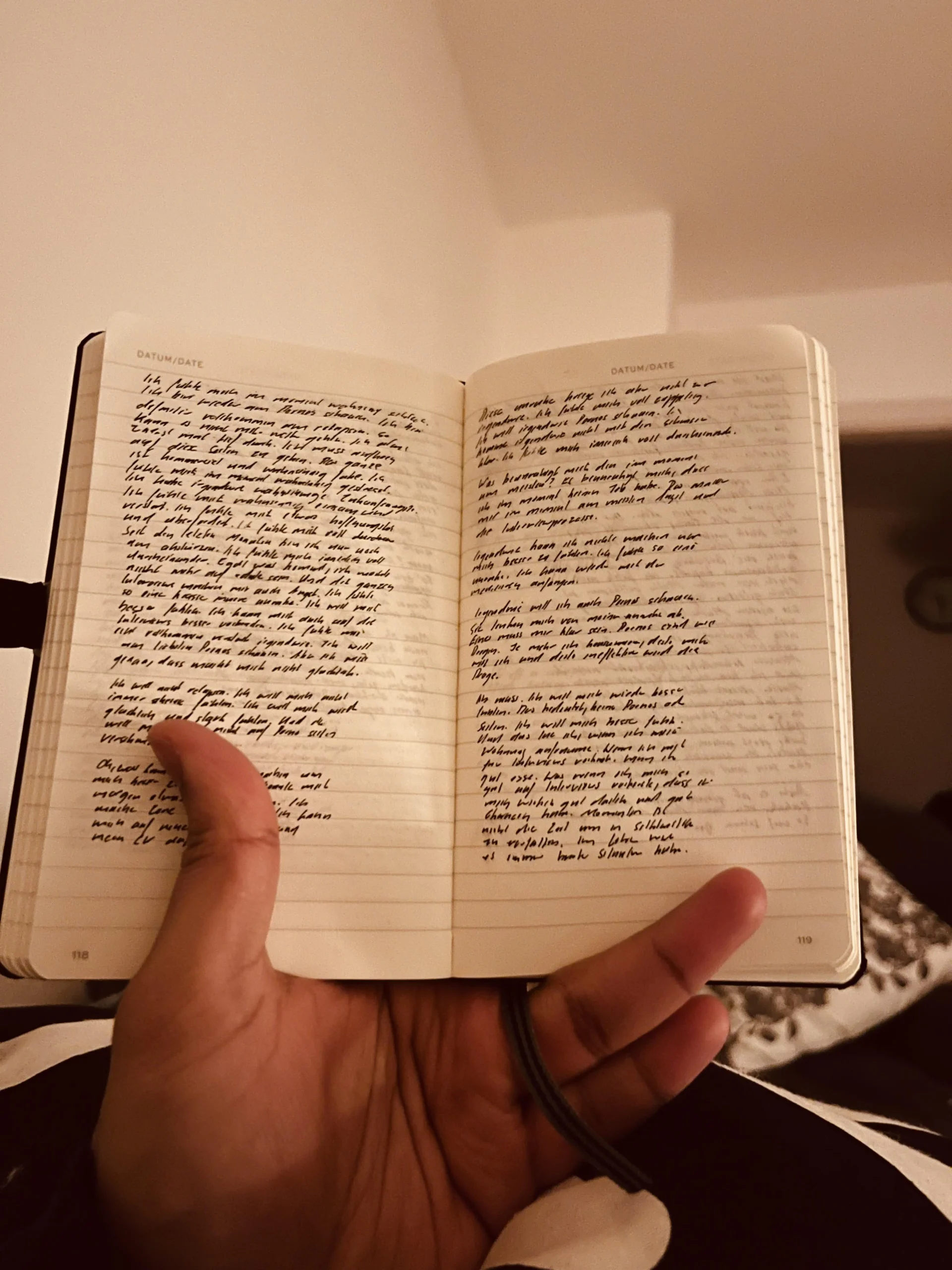 A hand holds open a small notebook filled with dense, handwritten text on both pages. The background is softly lit, showing part of a beige wall and ceiling. The writing appears personal, like journal entries.