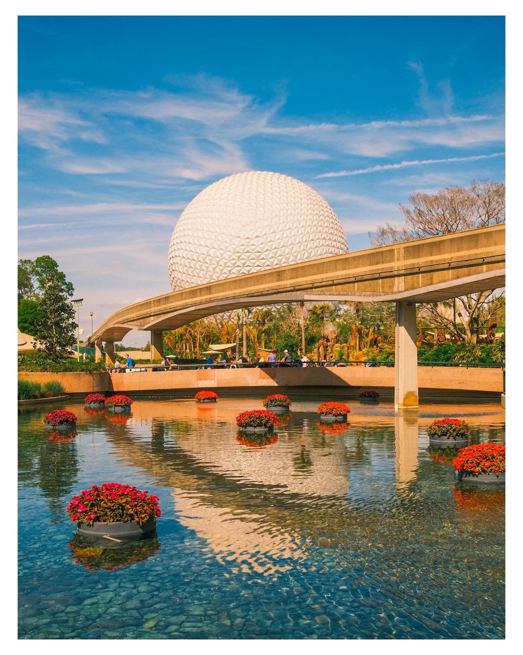 A white geodesic dome stands behind a monorail track and a reflective pond decorated with floating flower arrangements under a blue sky with light clouds.