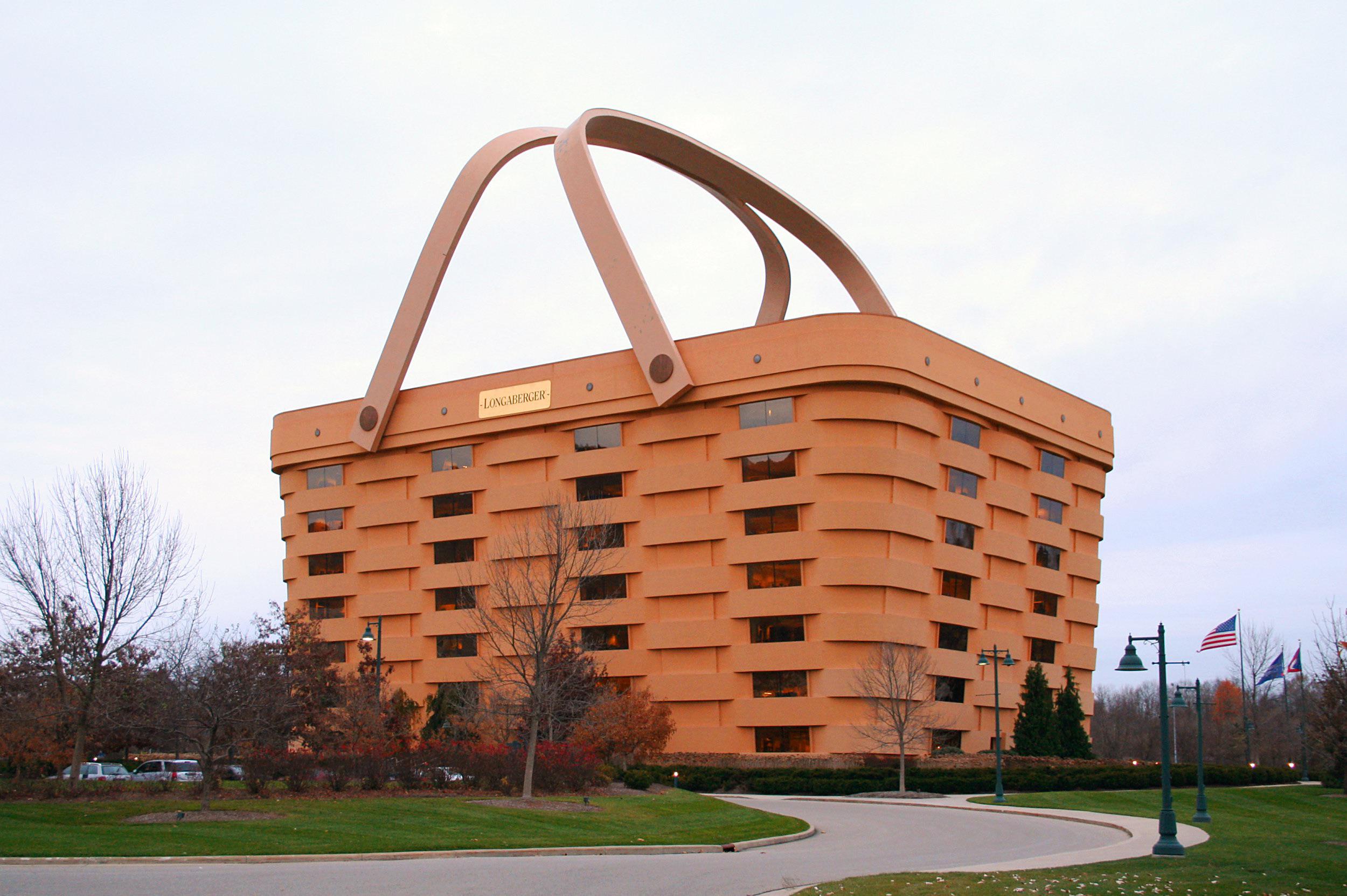 A large office building designed to look like a woven picnic basket with two giant handles, set in a landscaped area with trees, a road, streetlights, and American flags nearby.