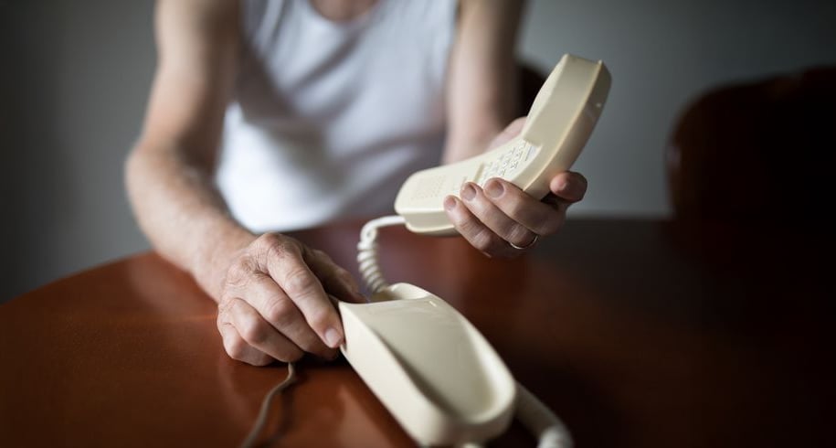 An older person in a sleeveless shirt sits at a table, holding the receiver of a beige corded telephone with one hand while resting the other hand on the base.
