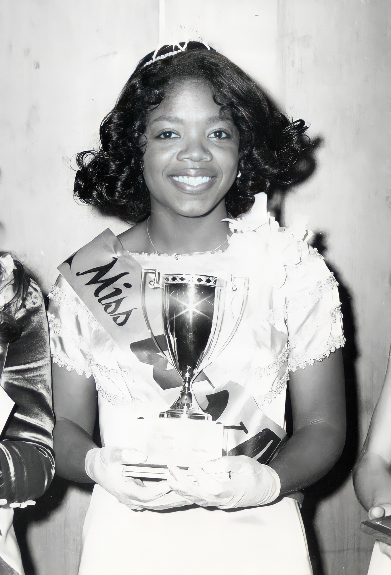 A smiling young woman wearing a crown and sash holds a trophy. She is dressed in a formal gown with gloves and a corsage, standing in front of a neutral background.