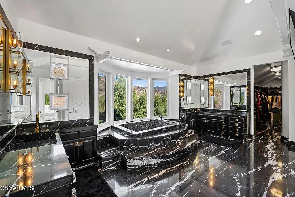 Luxurious black marble bathroom with a large soaking tub centered by bay windows, surrounded by double vanities, mirrored walls, gold accents, and a spacious walk-in closet in the background.