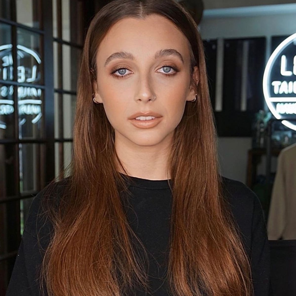 A young woman with long, straight brown hair and blue eyes is standing indoors, wearing a black shirt. She is looking directly at the camera with a neutral expression. The background is softly lit with glass panels and a neon sign.