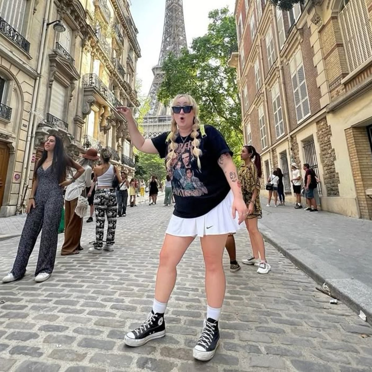 A woman with blonde braids, sunglasses, and a graphic t-shirt poses energetically on a cobblestone street in front of the Eiffel Tower, surrounded by several other people.
