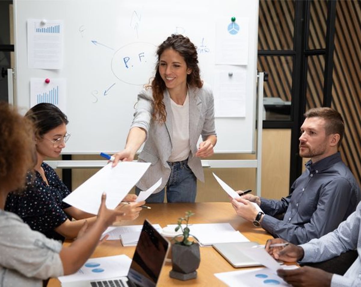 A woman stands and smiles while handing out papers to colleagues seated around a conference table in a meeting room. A whiteboard with diagrams and notes is in the background.