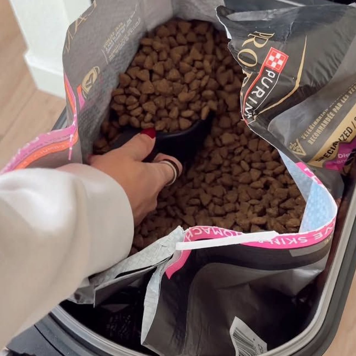 A hand holding a scoop is reaching into an open bag of Purina dry pet food, with kibble visible inside the bag. The bag is placed inside a larger container.