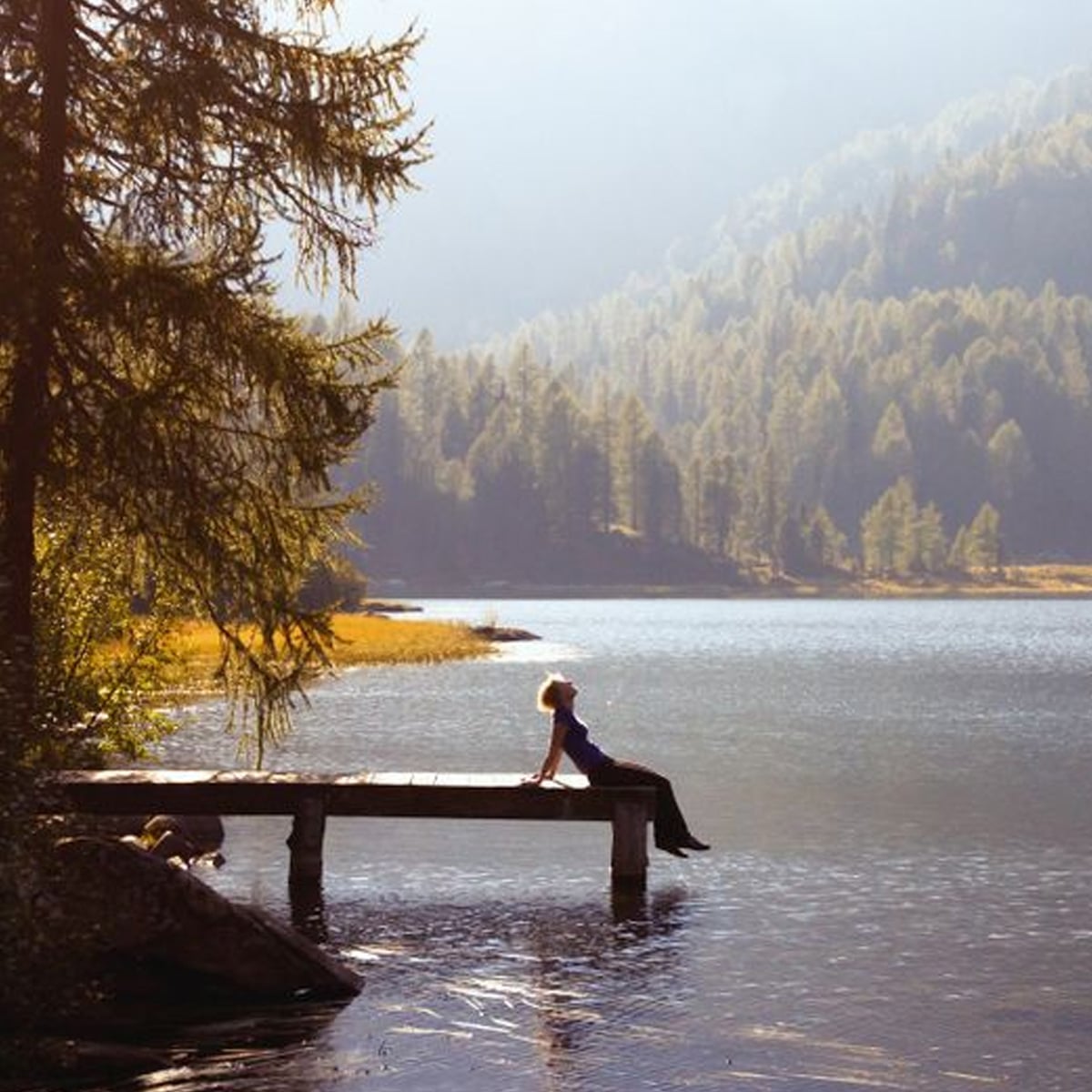 A person sits relaxing on a wooden dock by a calm lake, surrounded by tall pine trees and misty forested mountains in the background, with sunlight filtering through the scene.
