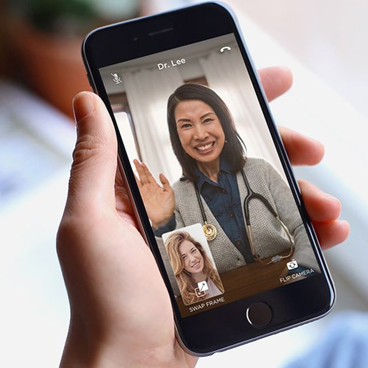 A person holds a smartphone showing a video call with a smiling female doctor wearing a stethoscope and waving. The screen also displays a small window with the caller’s face and camera options.