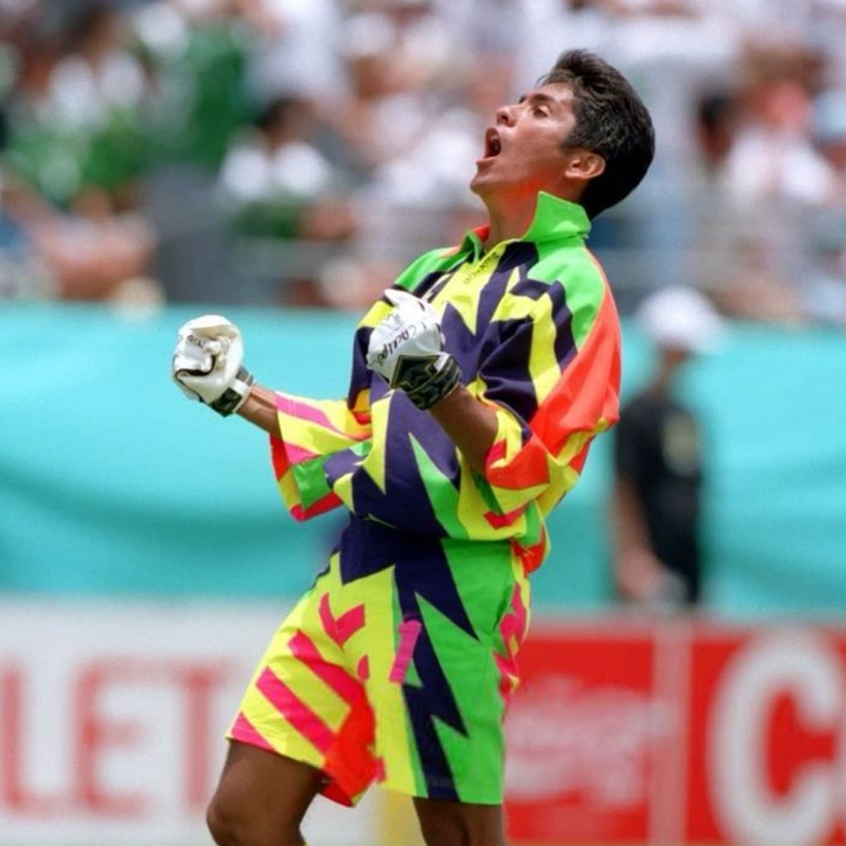 A cricket player wearing a brightly colored, neon zigzag-patterned uniform celebrates passionately on the field, clenching fists and shouting, with an audience blurred in the background.