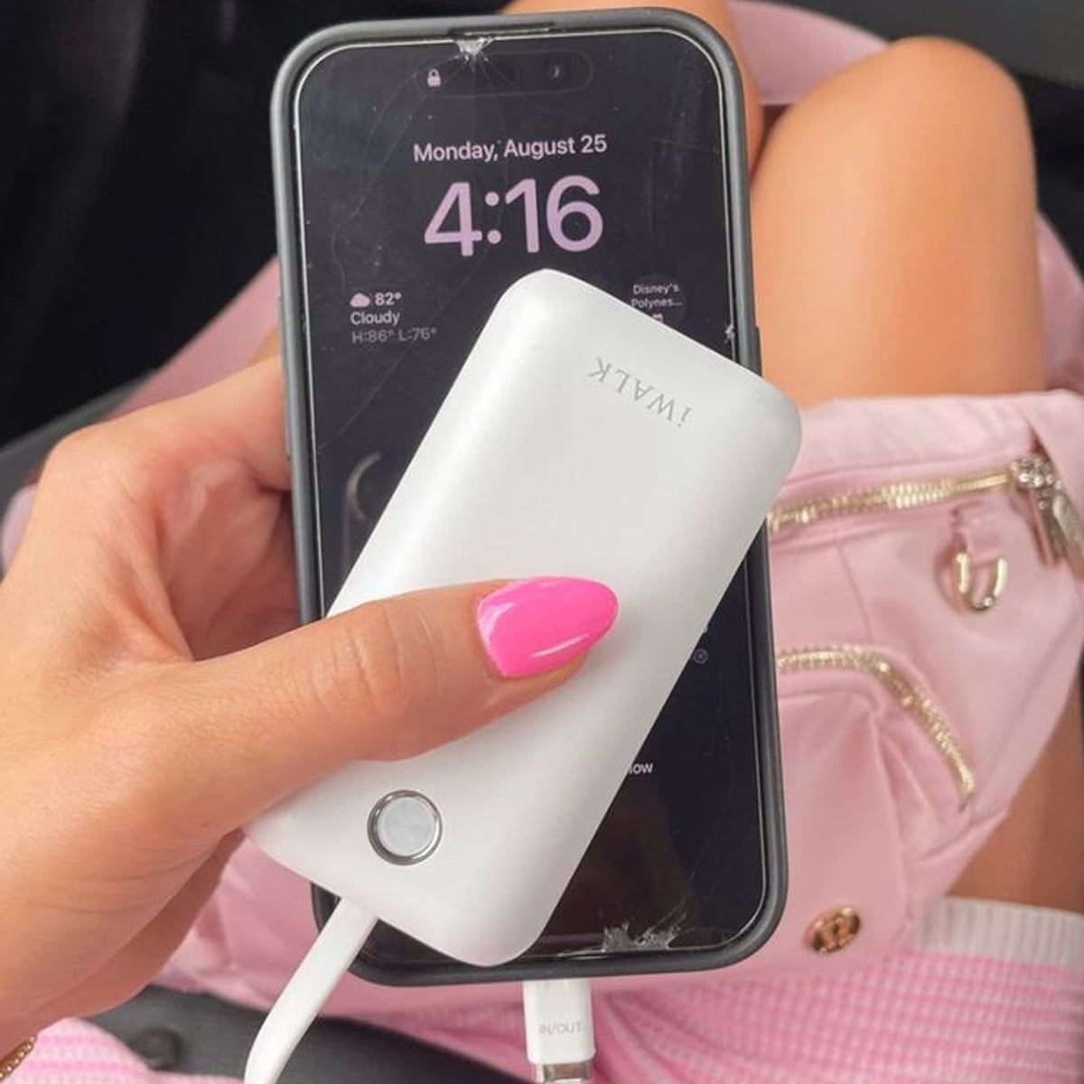A person with pink nail polish holds a white iWALK power bank charging a black phone displaying 4:16 on August 25. A pink bag with a zipper is in the background.