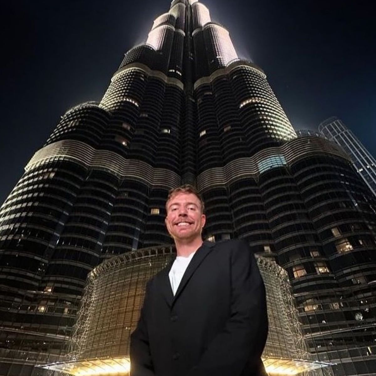 A man in a black suit smiles while standing in front of the illuminated Burj Khalifa skyscraper at night. The building towers above, glowing with lights against the dark sky.