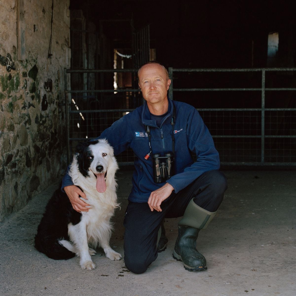 A man in outdoor clothing and boots kneels beside a black and white dog, resting his hand on the dog's back. They are in a barn with stone walls and metal gates in the background. The dog is sitting and panting.