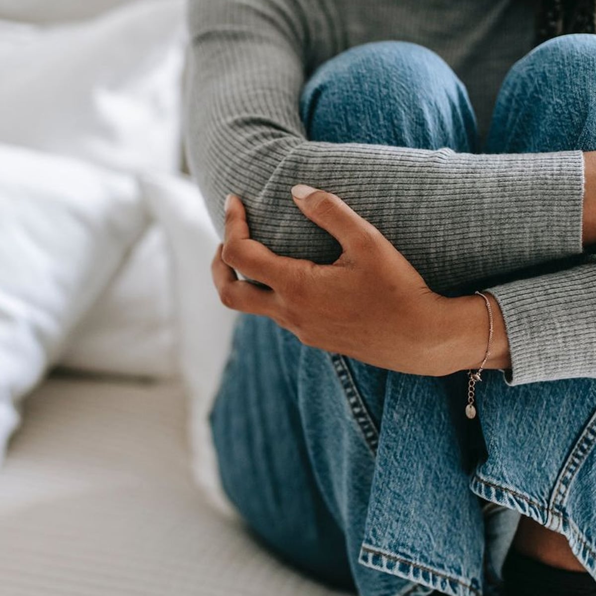 A person wearing a gray ribbed sweater and blue jeans sits on a bed with knees pulled to their chest, arms wrapped around their legs, and a silver bracelet on their wrist. White bedding is visible in the background.