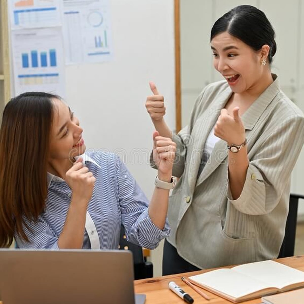Two women in an office setting smile and give each other thumbs up. One is seated at a desk with a laptop and notebook, while the other stands beside her, both appearing cheerful and celebratory.