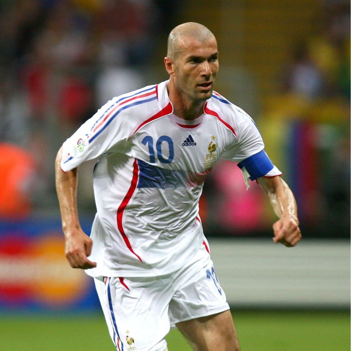 A male soccer player in a white jersey with blue and red stripes, number 10, runs on the field. He wears a blue captain’s armband and appears focused during a match. The background is blurred with stadium lights and spectators.