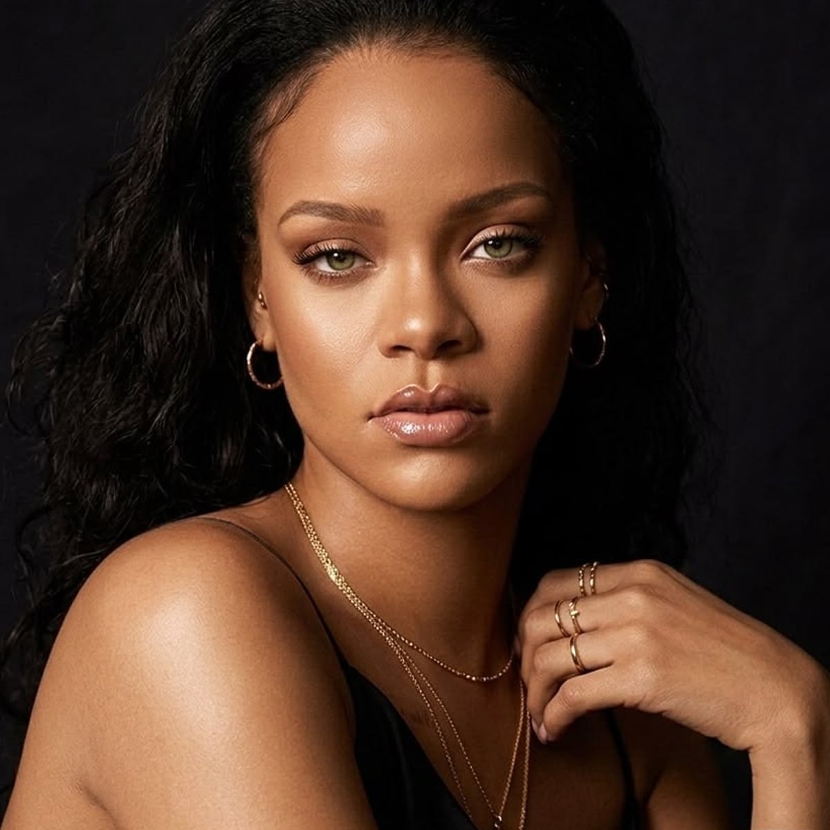 A woman with long dark curly hair and light eyes gazes at the camera. She wears hoop earrings, layered gold necklaces, stacked rings, and a black top, with a dark background behind her.