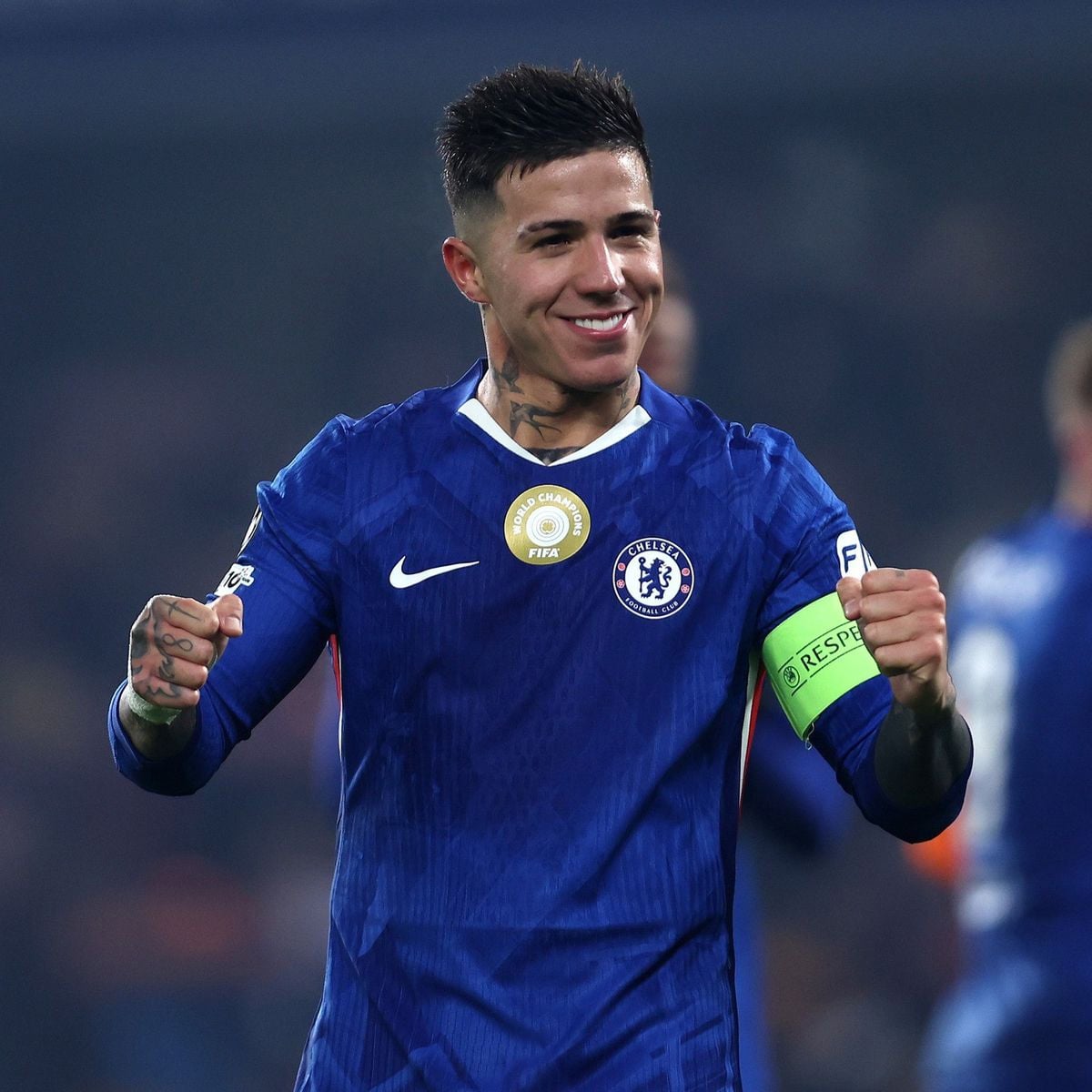 A male soccer player wearing a blue Chelsea FC jersey with a green captain’s armband smiles and raises his fists in celebration on the field.