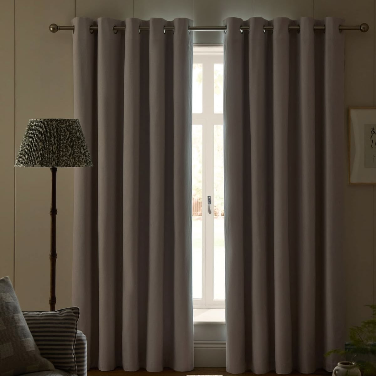 A window with closed, light gray curtains hanging from a rod. A floor lamp with a patterned shade stands to the left, and part of a couch with striped cushions is visible in the foreground.