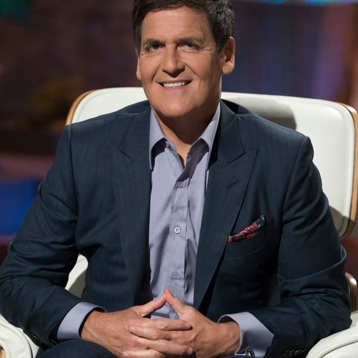 A man in a dark suit and light shirt sits smiling on a white chair, hands clasped, in a well-lit studio setting with a blurred background.