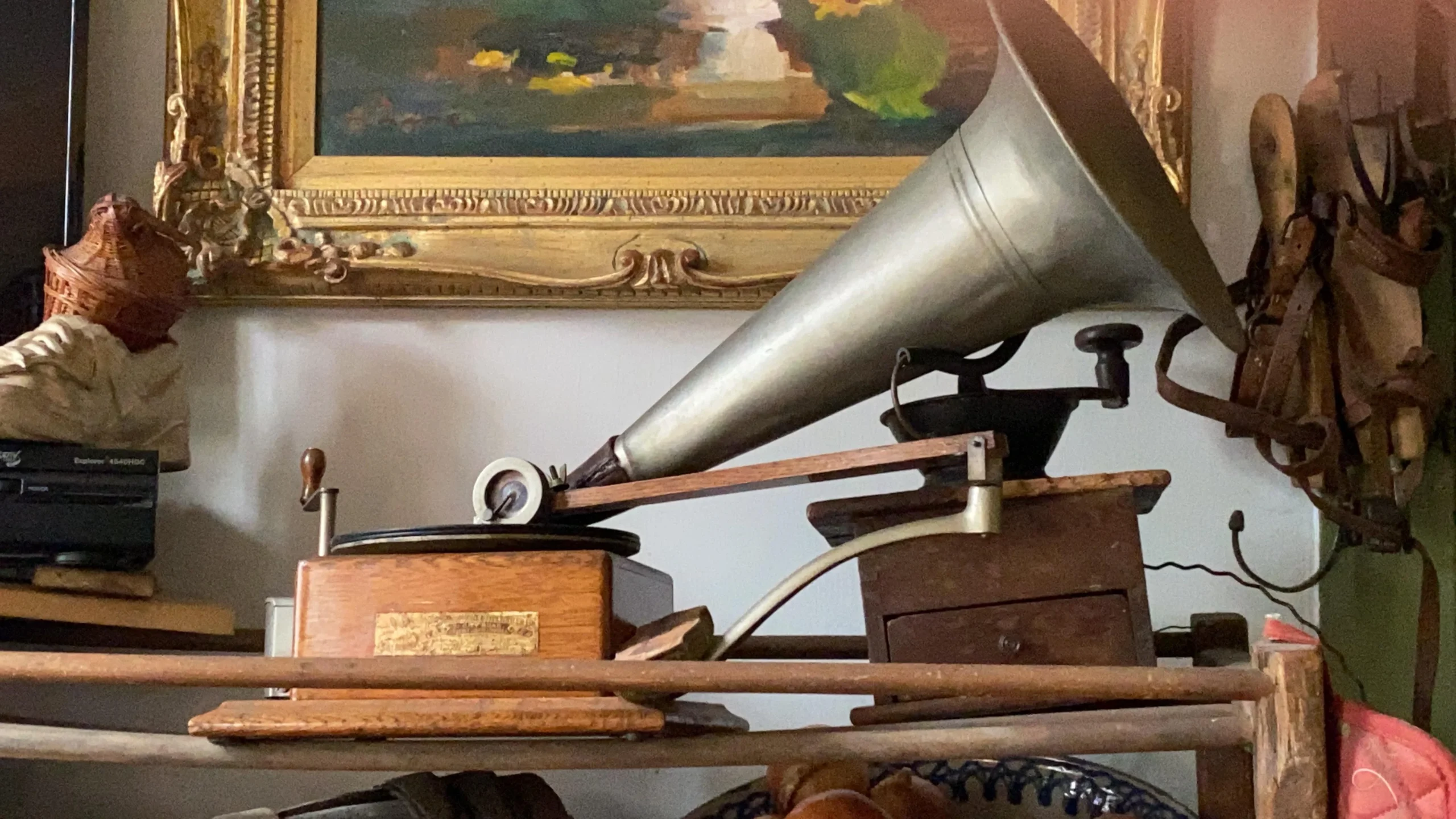 A close-up of an antique gramophone with a large horn speaker, wooden base, and metal handle, displayed indoors with a framed painting and various vintage items in the background.