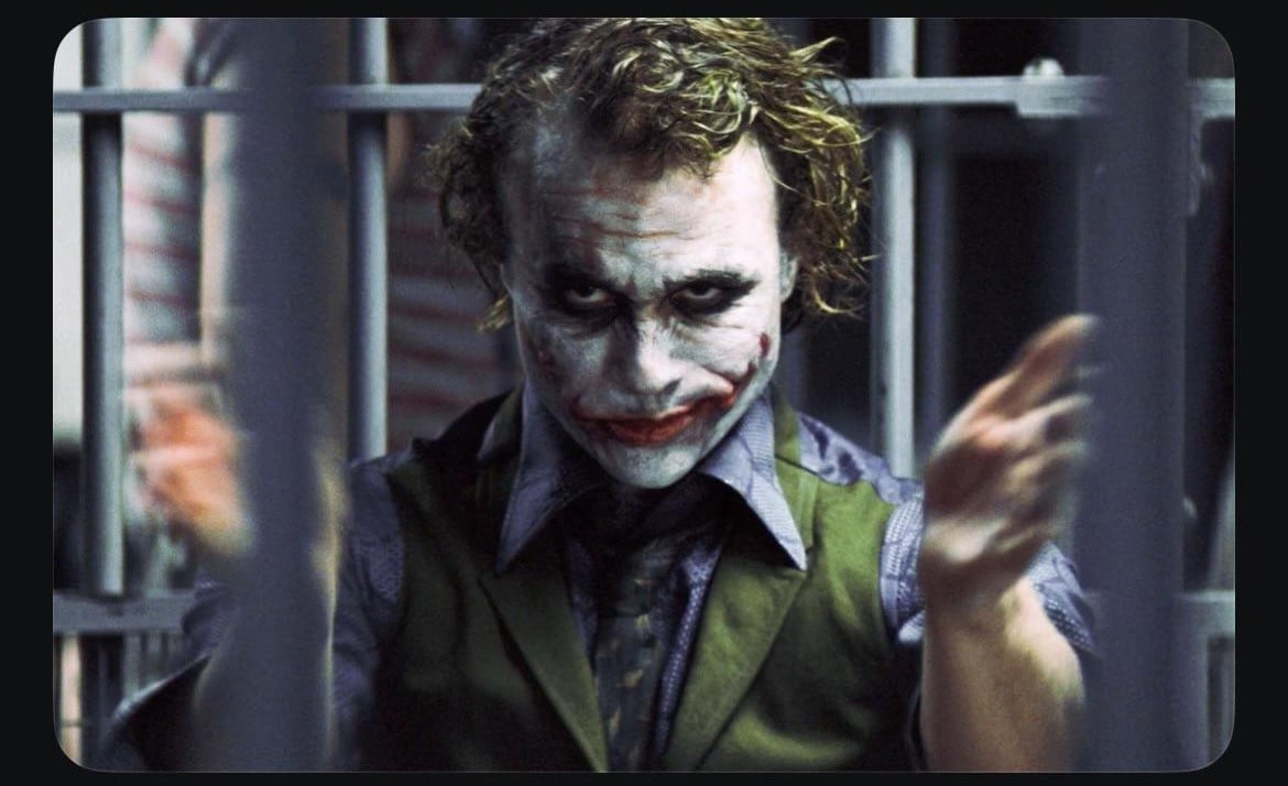 A person with messy hair and smeared face paint sits behind jail bars, wearing a vest and collared shirt, with hands raised and a menacing expression highlighted by dark eye makeup and a red, exaggerated mouth.