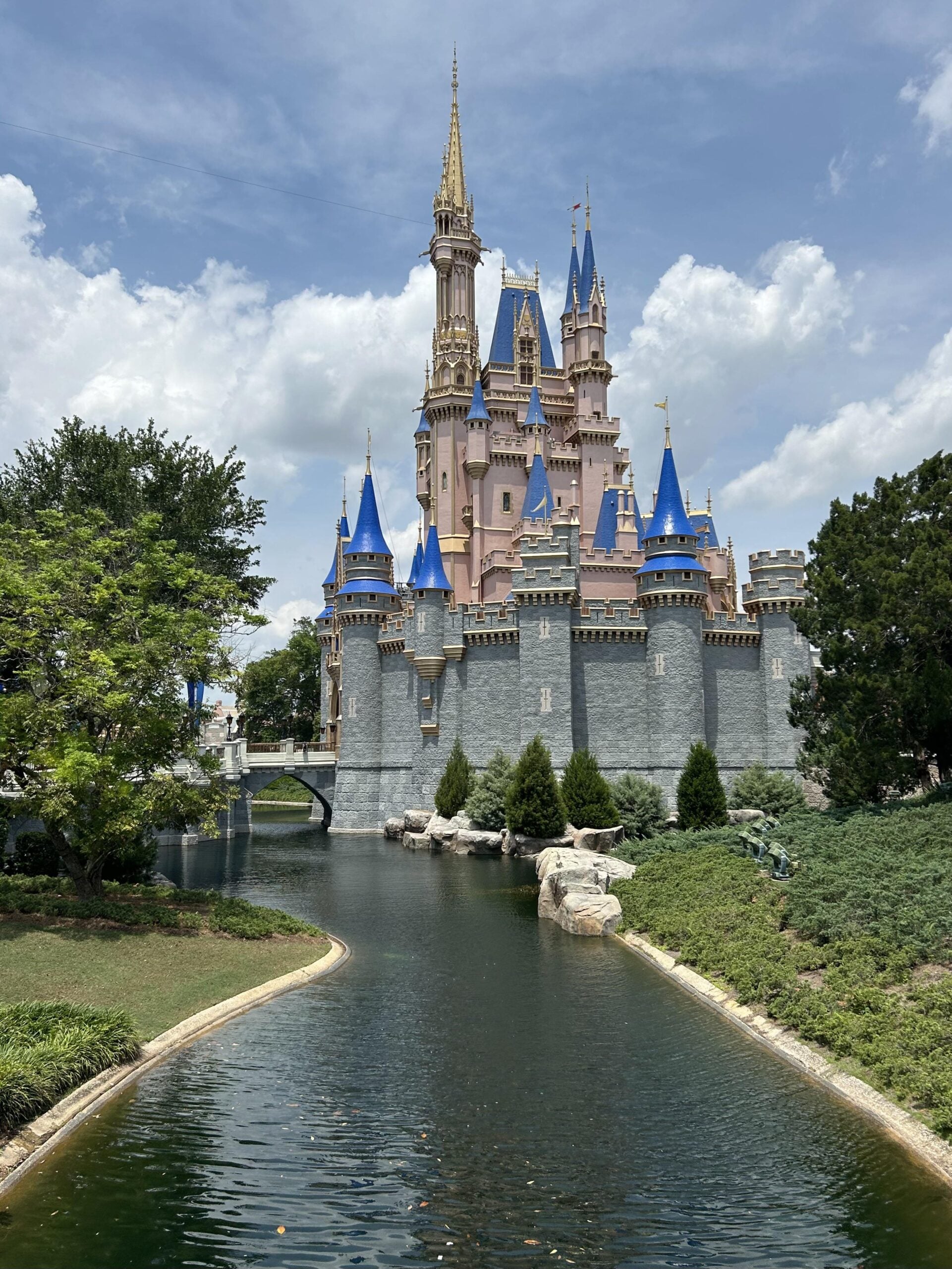 A large fairytale castle with pink walls, blue rooftops, and gold spires stands beside a moat, surrounded by greenery and trees under a partly cloudy sky.