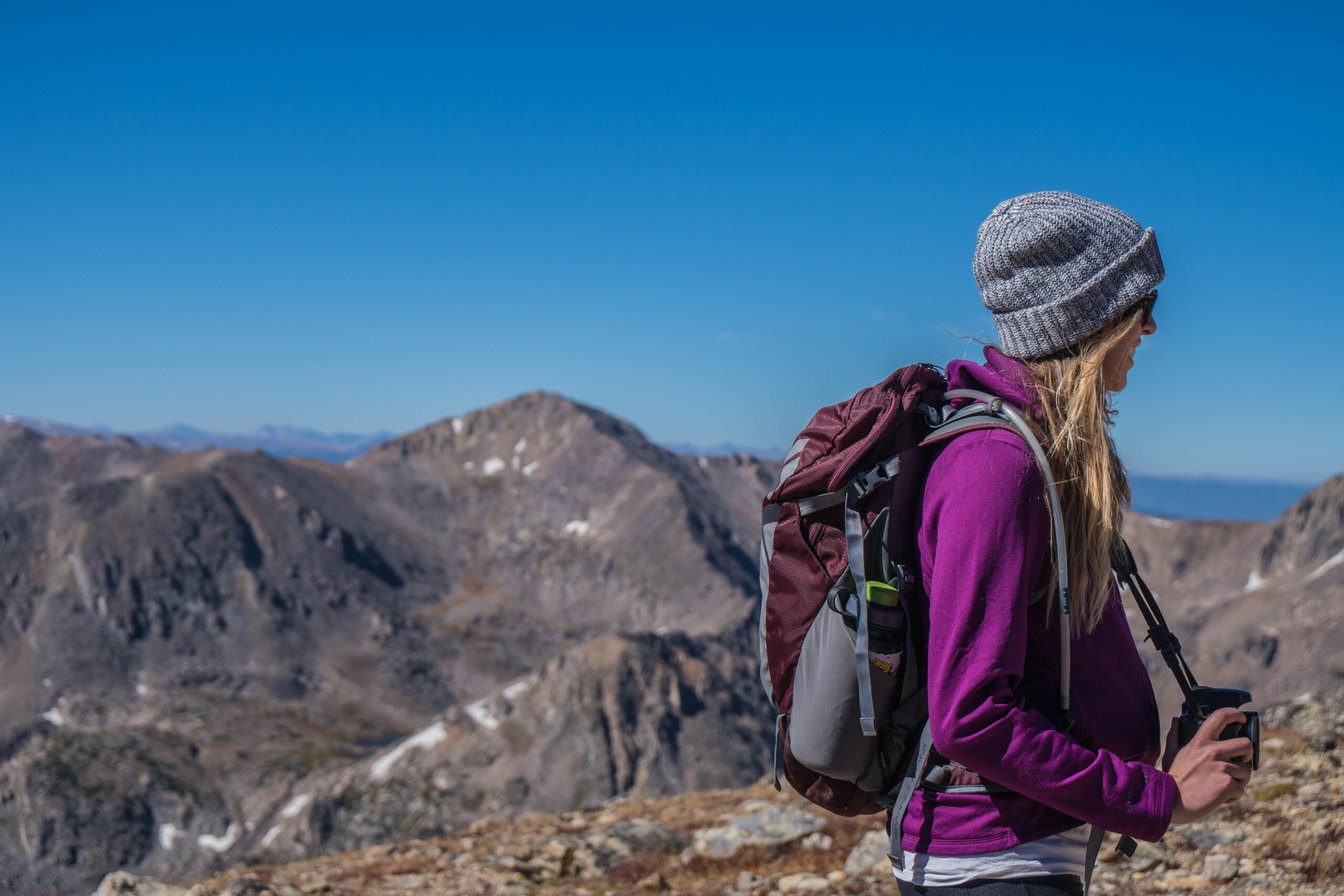 A woman in a gray beanie and purple jacket with a backpack stands on a rocky mountain, gazing at the distant peaks under a clear blue sky.
