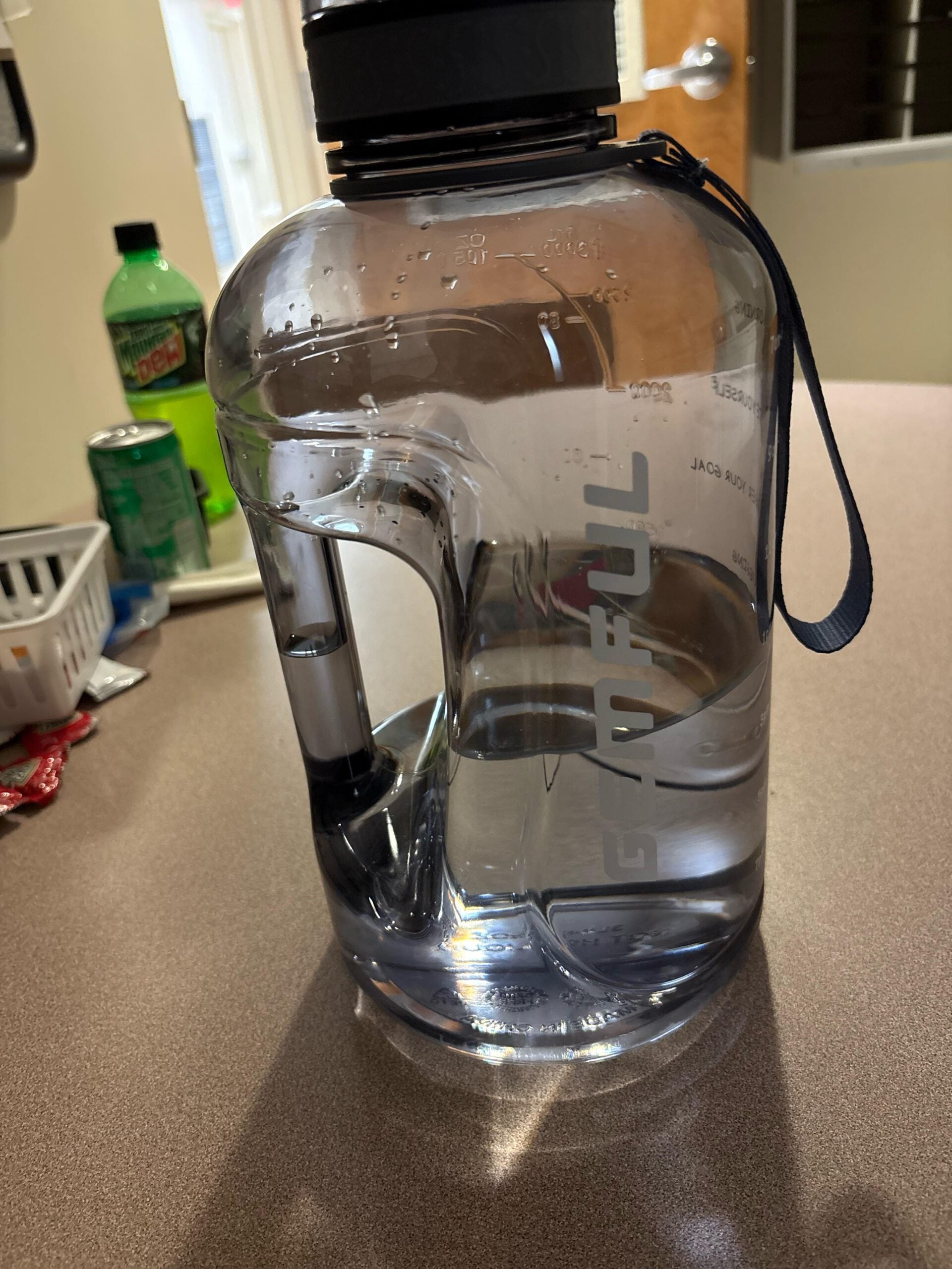 A large, clear water jug with a black lid and a carrying strap sits on a countertop. In the background are a green Mountain Dew bottle, some snacks, and a partially open door.