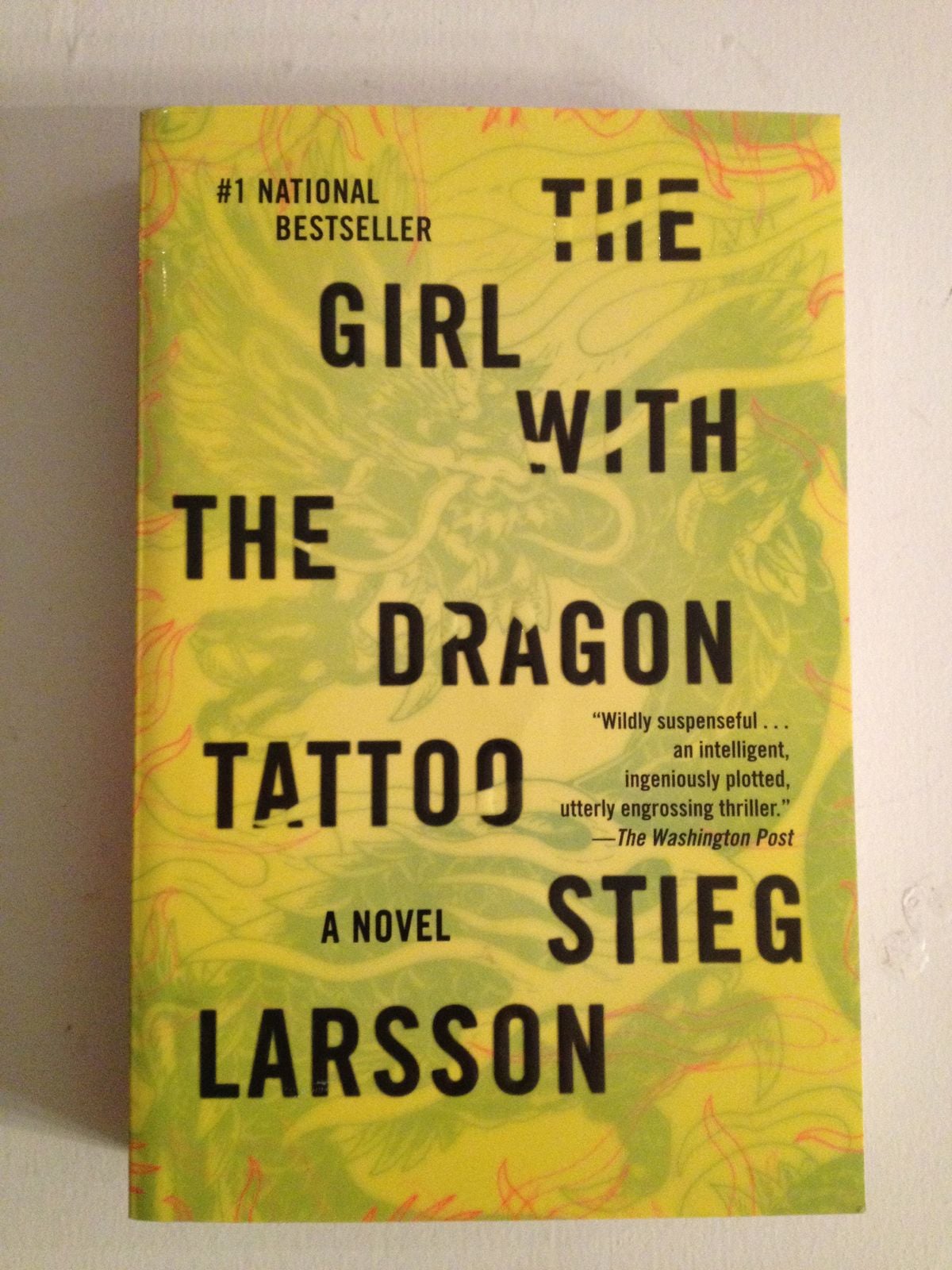 The cover of the book "The Girl with the Dragon Tattoo" by Stieg Larsson, featuring bold black text on a yellow background with a faint dragon illustration.