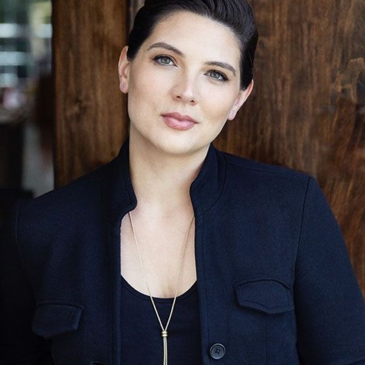 A woman with fair skin and dark hair pulled back, wearing a black jacket over a black top, stands in front of a wooden wall and looks confidently at the camera. She has a long necklace and a calm expression.