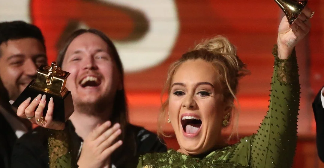 A woman in a green dress holds up a Grammy award, smiling joyfully, with people clapping and cheering in the background.