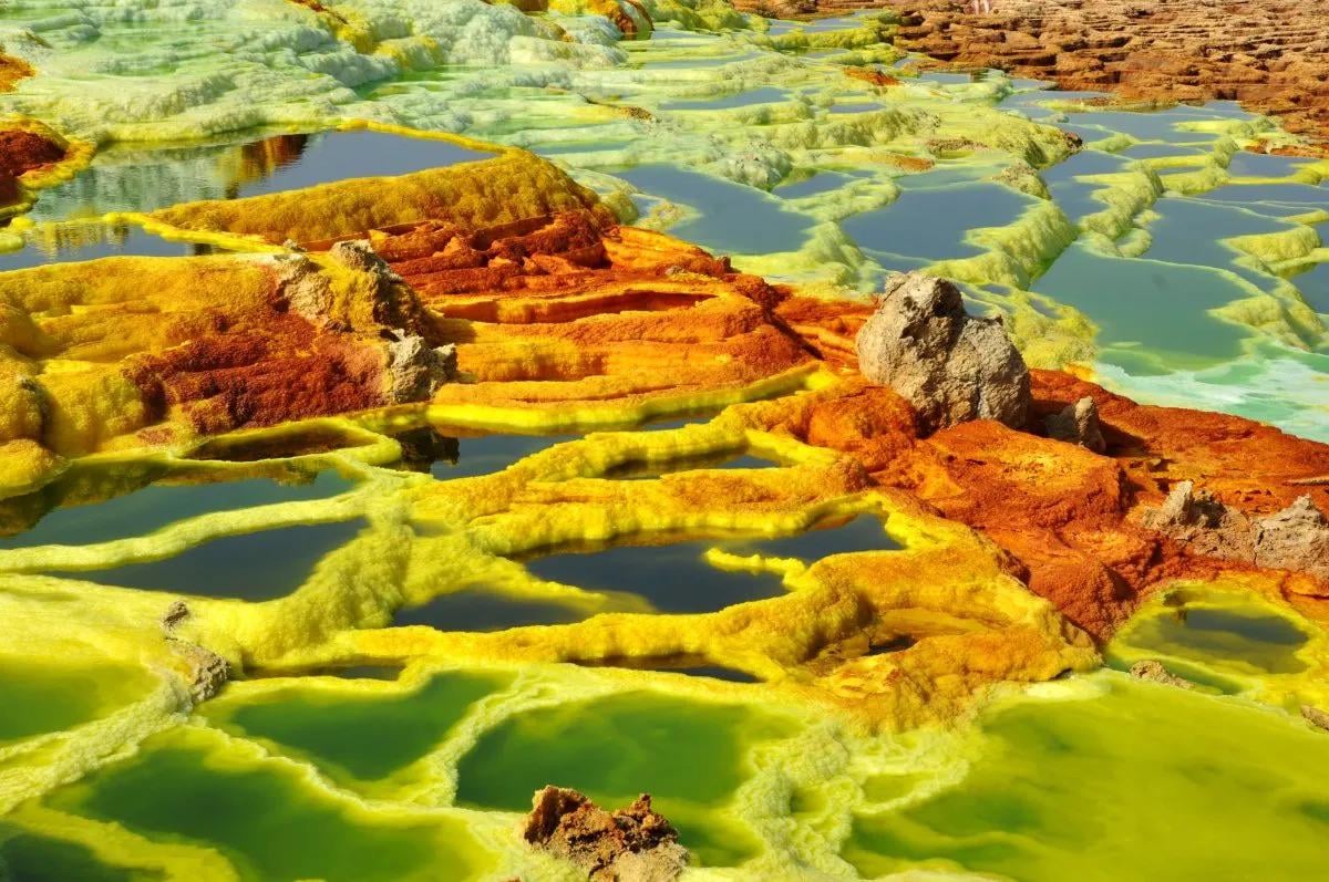 A vibrant geothermal landscape with pools of green and yellow water, separated by orange and yellow mineral deposits and rocky formations.