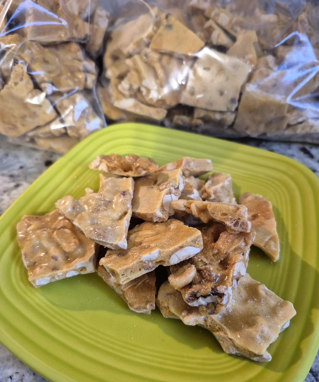 Pieces of peanut brittle are stacked on a green plate, with bags of additional peanut brittle in the background on a countertop.