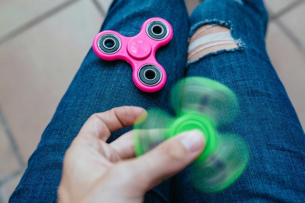 A person wearing ripped blue jeans holds and spins a green fidget spinner, while a stationary pink fidget spinner rests on their lap.