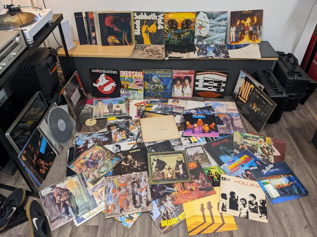 A large collection of vinyl records spread across the floor, with many colorful album covers visible, and more records displayed upright on a bench behind them. A turntable is partially visible on the left.