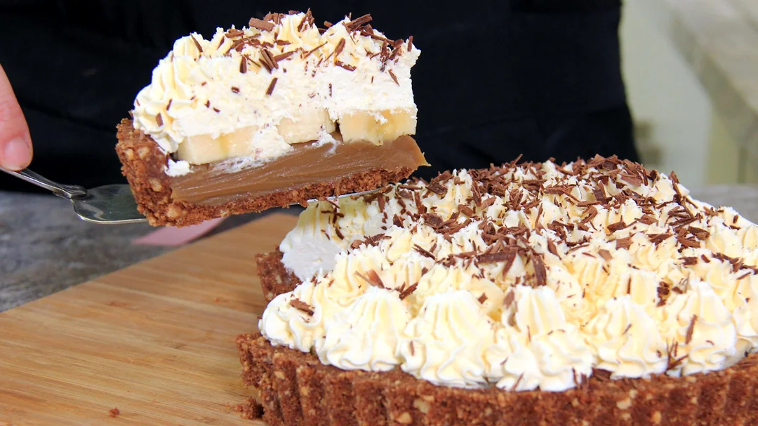 A hand lifts a slice of banoffee pie topped with whipped cream and chocolate shavings, revealing layers of banana and caramel filling in a crumbly crust, with the remaining pie on a wooden board.