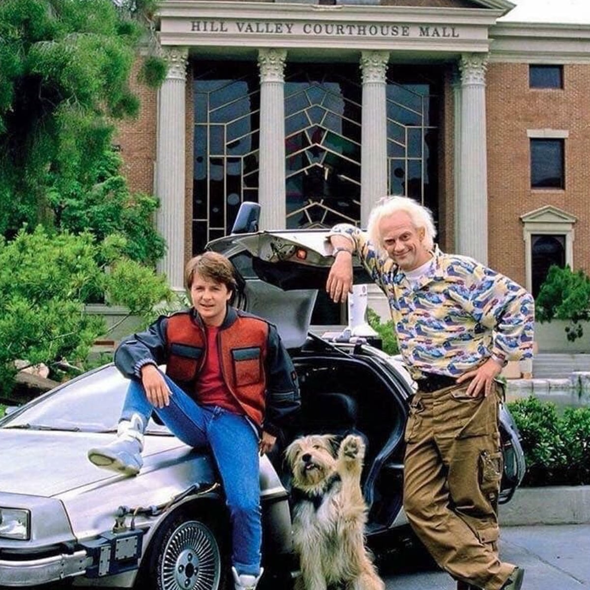 Two men and a dog pose in front of a DeLorean car with gull-wing doors open, outside the Hill Valley Courthouse. The younger man sits on the car, while the older man stands, both smiling.