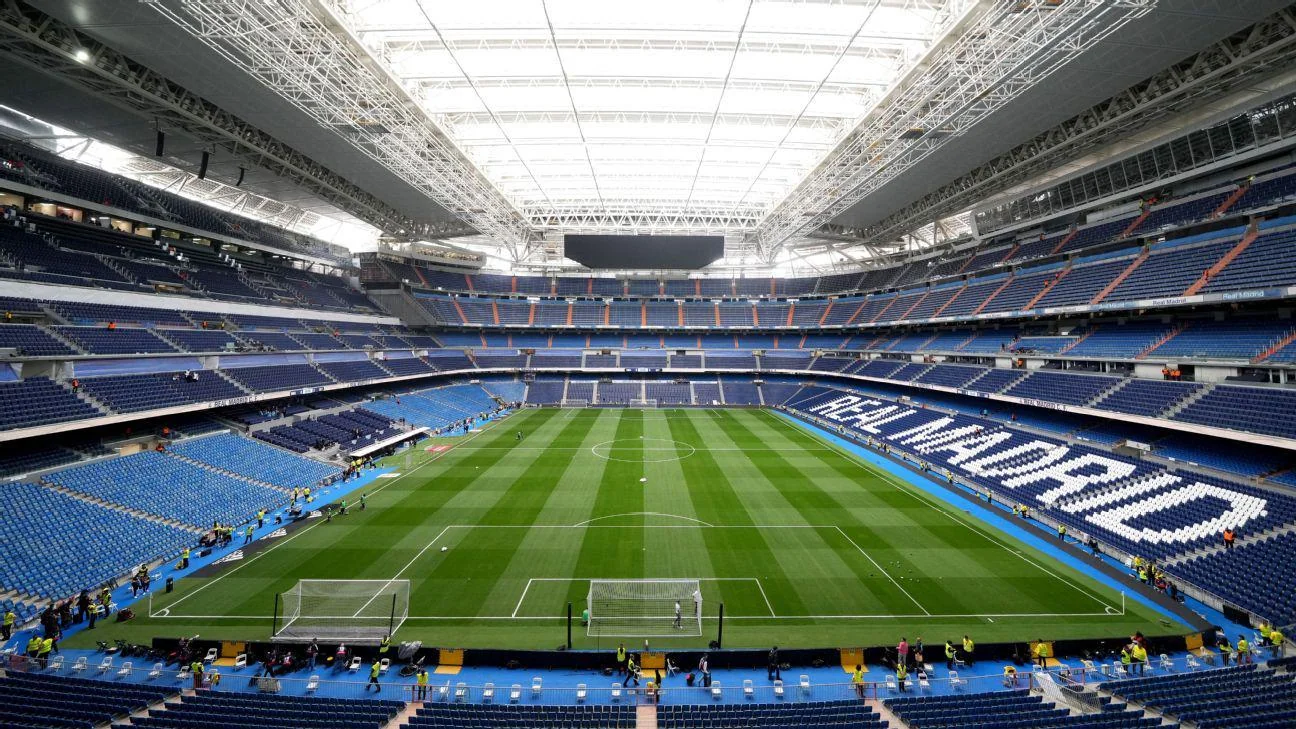 A wide view of a modern football stadium with a glass roof, green pitch, and blue seats. "REAL MADRID" is spelled out in white letters across the stands. The stadium is mostly empty with a few people near the pitch.