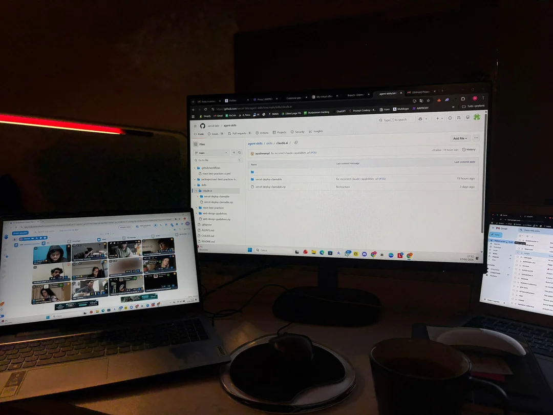 A dimly lit desk with three screens: a laptop showing a video call grid, a large monitor displaying a GitHub repository, and a smaller laptop showing file folders. A mouse, mousepad, and cup are also on the desk.