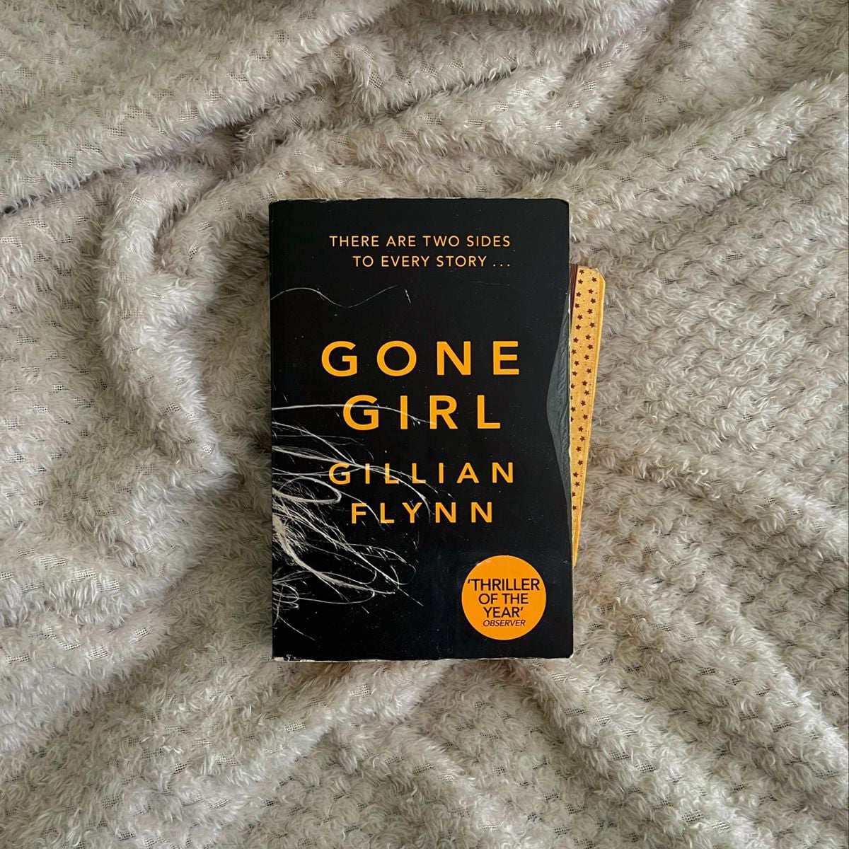 A copy of the book "Gone Girl" by Gillian Flynn rests on a textured, light gray blanket. The cover features bold yellow text and a sticker that reads "Thriller of the Year.