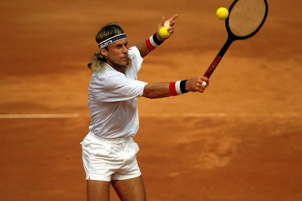 A tennis player wearing a white outfit and headband hits a two-handed backhand on a clay court, with a tennis ball in mid-air near the racquet.