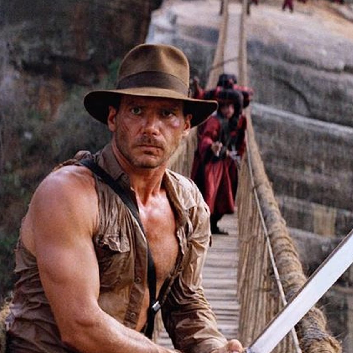 A man in a brown hat and rugged clothing stands on a rope bridge, holding a large sword. Behind him, people dressed in red are crossing the bridge against a rocky canyon backdrop.