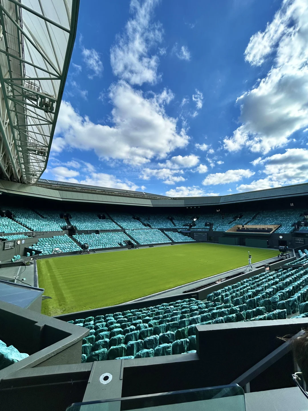 View of an empty tennis stadium under a partly cloudy sky, with teal-colored seats surrounding a grass court. Some maintenance staff are visible on the court and near the stands.