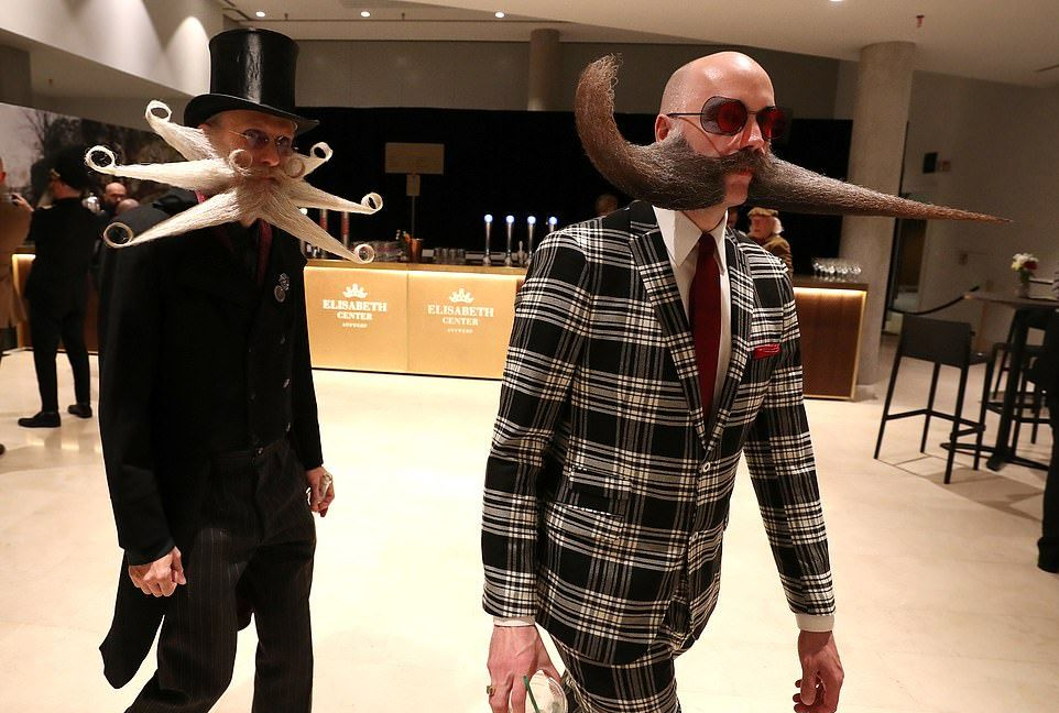 Two men with extravagant, sculpted mustaches walk indoors. One wears a tall hat and black suit, the other wears a plaid suit and large sunglasses. Both have unusually large and styled facial hair.